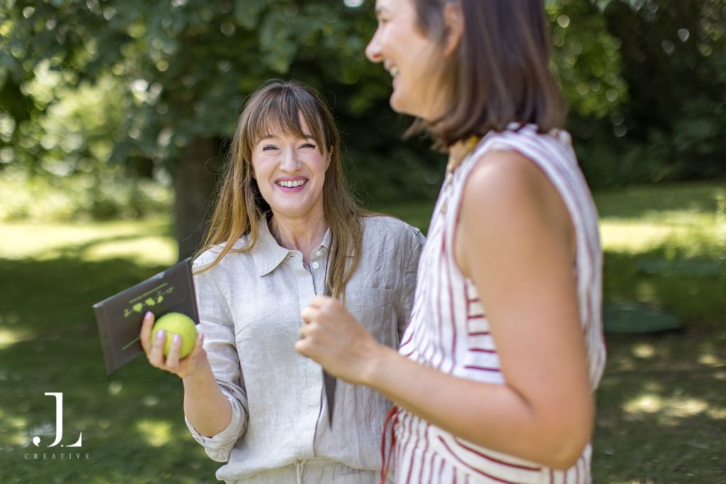 Candid outdoor photo of a woman smiling while holding brochures and a green ball in a garden