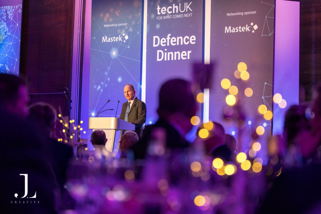 Speaker on stage with TechUK and sponsor branding at a London event