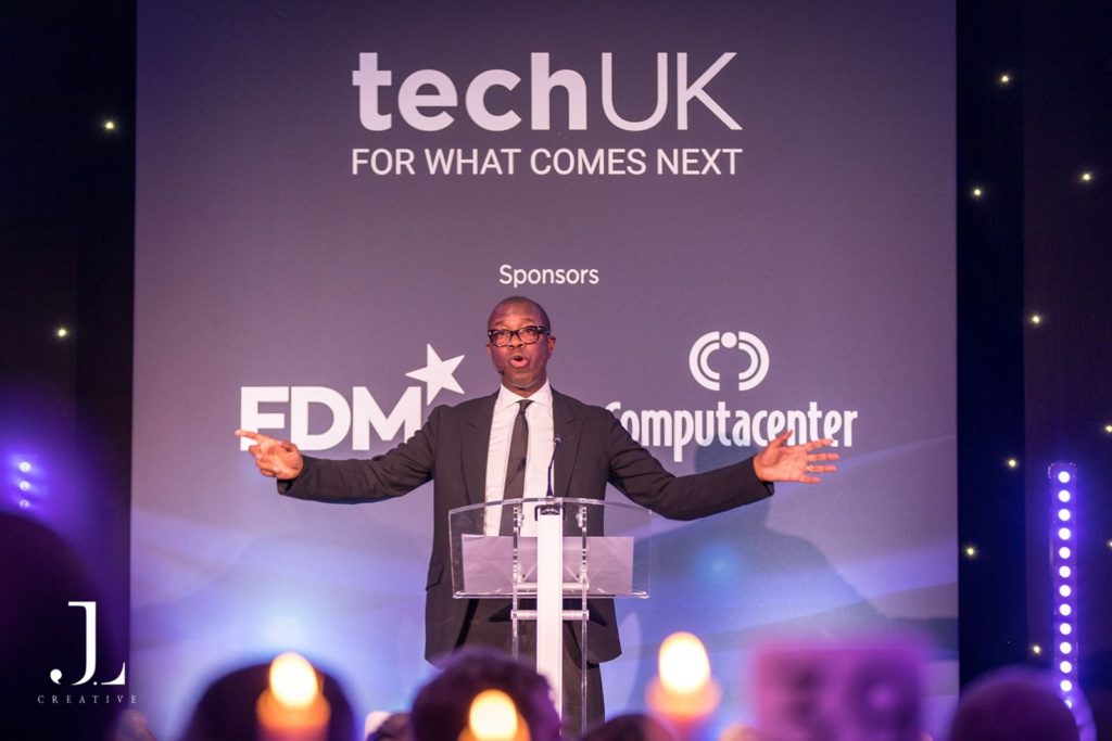 Speaker on stage with TechUK and sponsor branding at a London event