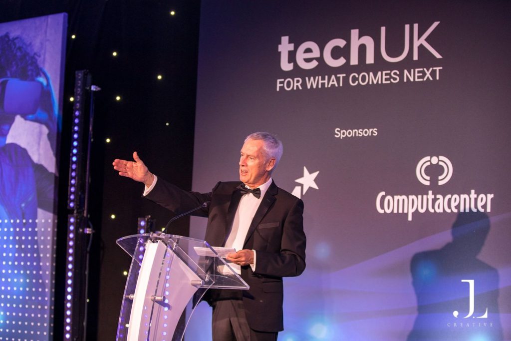 Speaker on stage with TechUK and sponsor branding at a London event