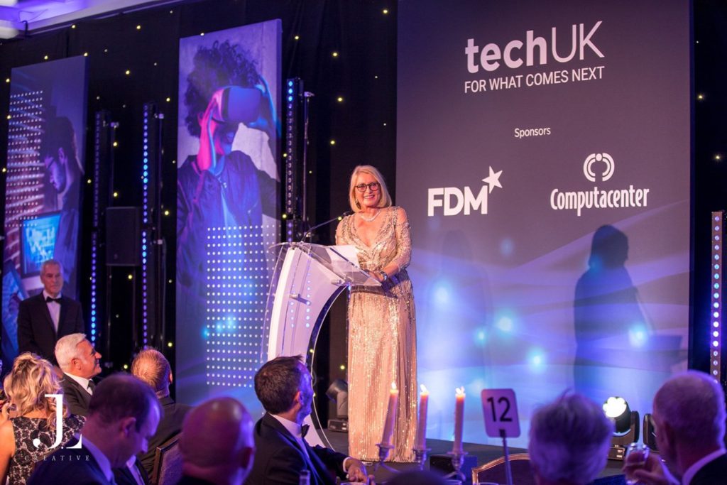 Speaker on stage with TechUK and sponsor branding at a London event