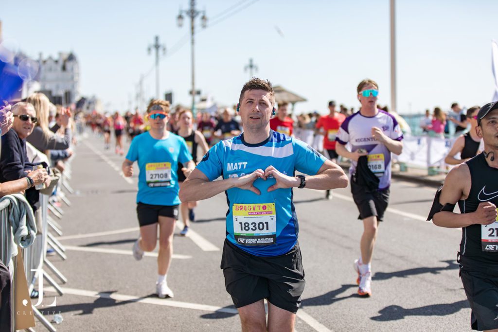 A male runner in a blue jersey making a heart gesture with his hands during the Brighton Marathon.