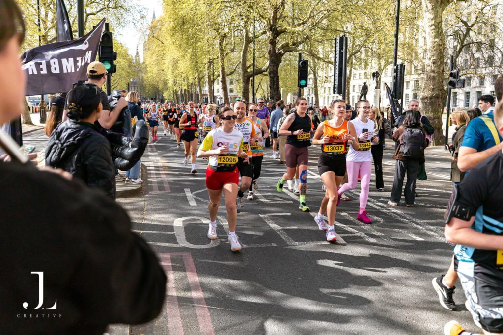 A crowd of diverse charity runners sprinting down a sunny, tree-lined London street