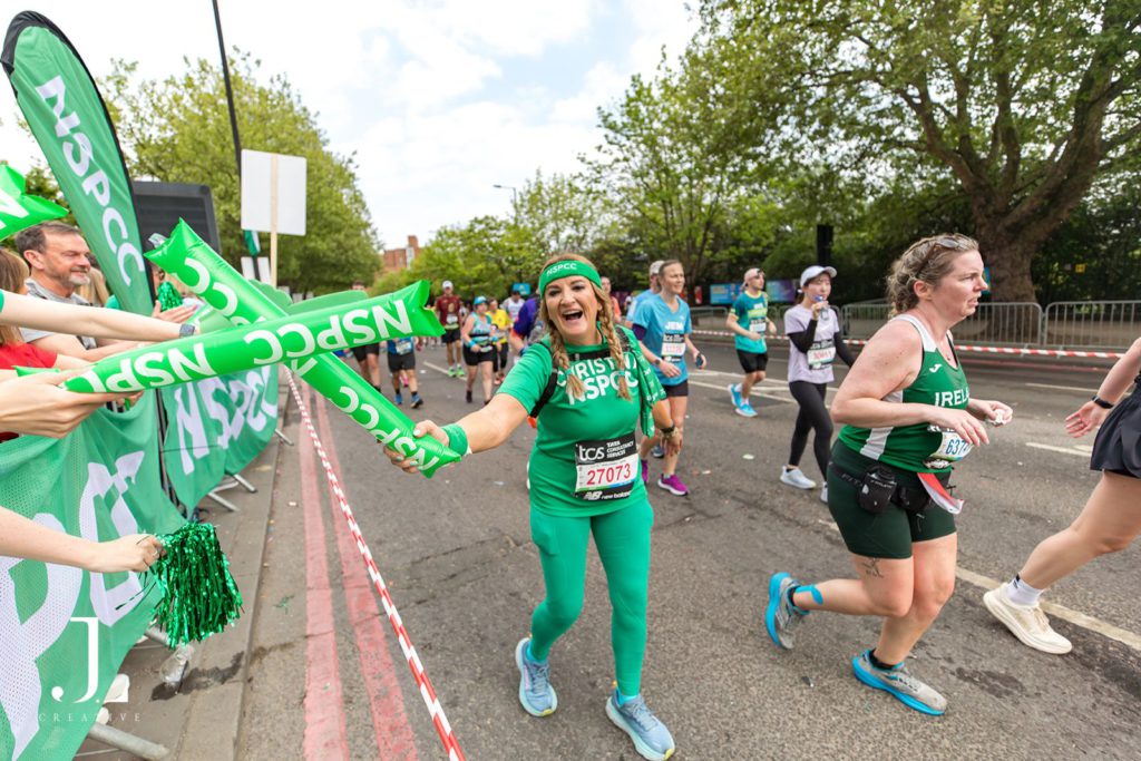 A joyful NSPCC runner high-fives supporters with green thundersticks at a marathon cheer point.