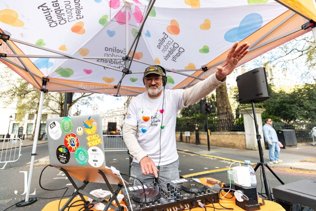 A DJ under an Evelina London Children’s Charity tent hyping up runners at a marathon cheer point.