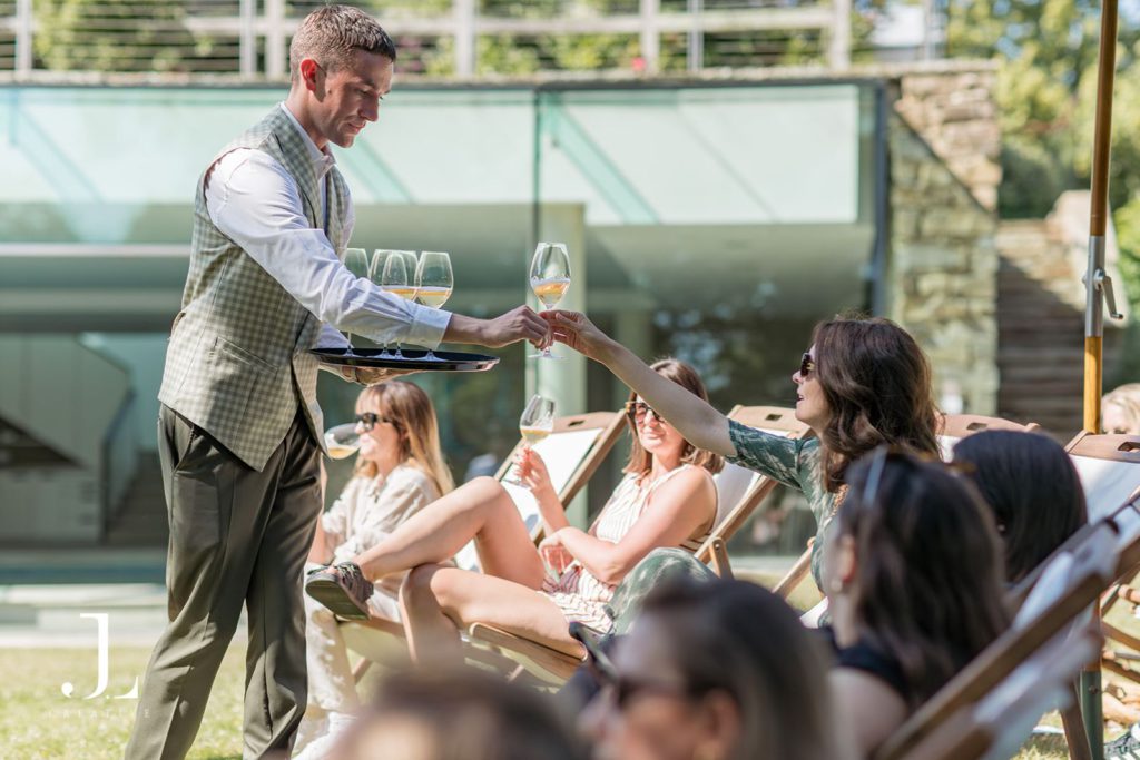 Luxury hospitality photography captures a waiter serving wine to guests lounging in a sunny outdoor setting.